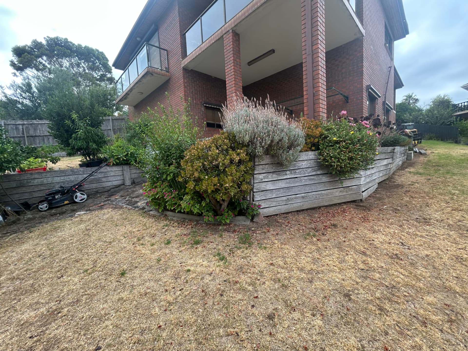 Aged timber sleeper garden wall and dry lawn beside a brick home before works