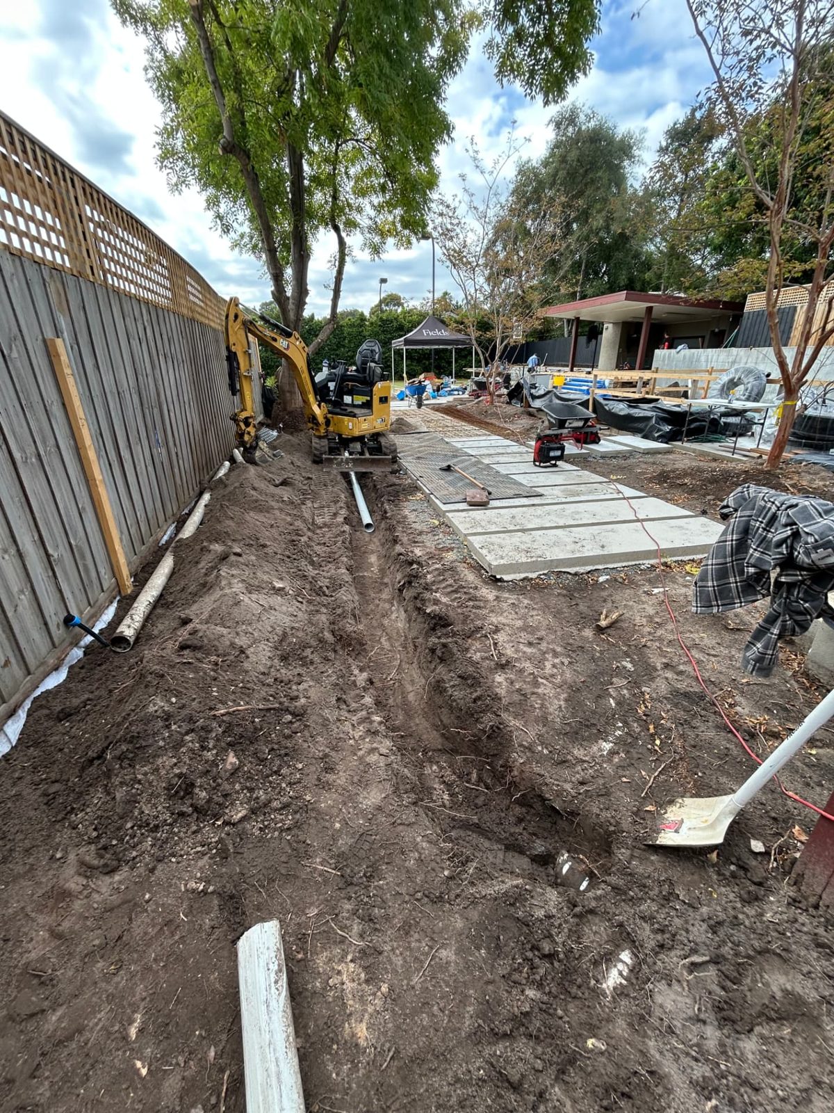Long drainage trench along a boundary fence with pipe laid out and new concrete pads beside it