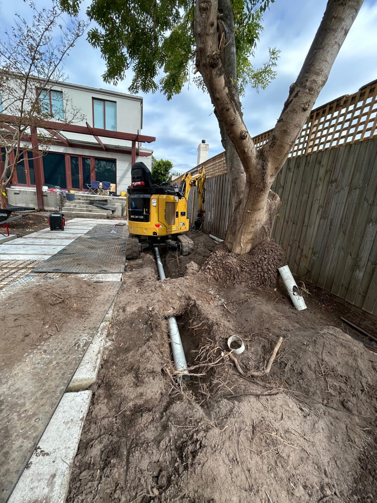 Branded Rigid Earthworks excavator laying stormwater pipe in a trench beside a large tree