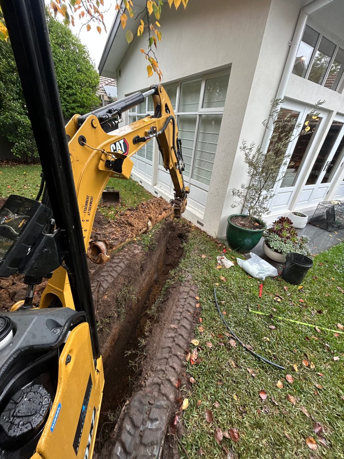 Compact Caterpillar excavator trenching along a lawn beside white timber windows